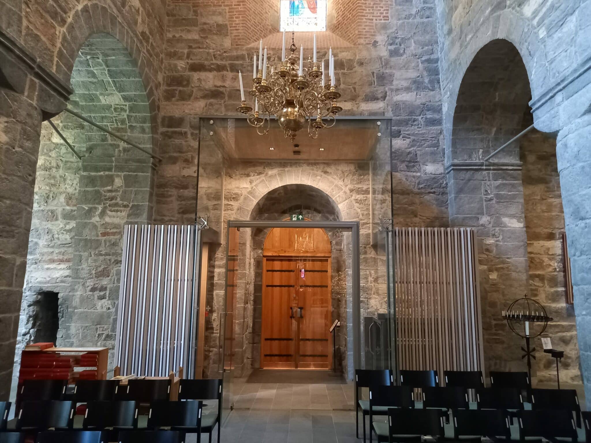 Interiør fra en middelalderkirke i stein. Bildet viser en inngangsport i tre rammet inn av en moderne glasskonstruksjon og vertikale trelameller. En stor messingkrone med stearinlys henger fra det hvelvede taket.