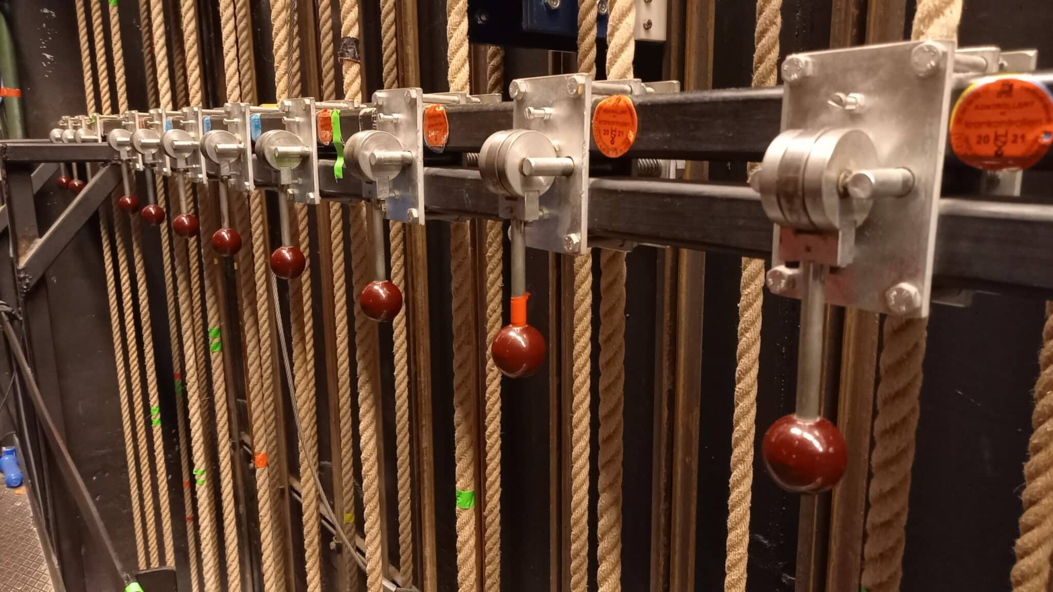 Nærbilde av et manuelt snortrekk-system i et teater. En rekke metallklemmer med brune, kuleformede håndtak er montert på en horisontal skinne. Tykke hampetau løper vertikalt gjennom systemet, og flere av klemmene har oransje kontrollmerker for året 2021.