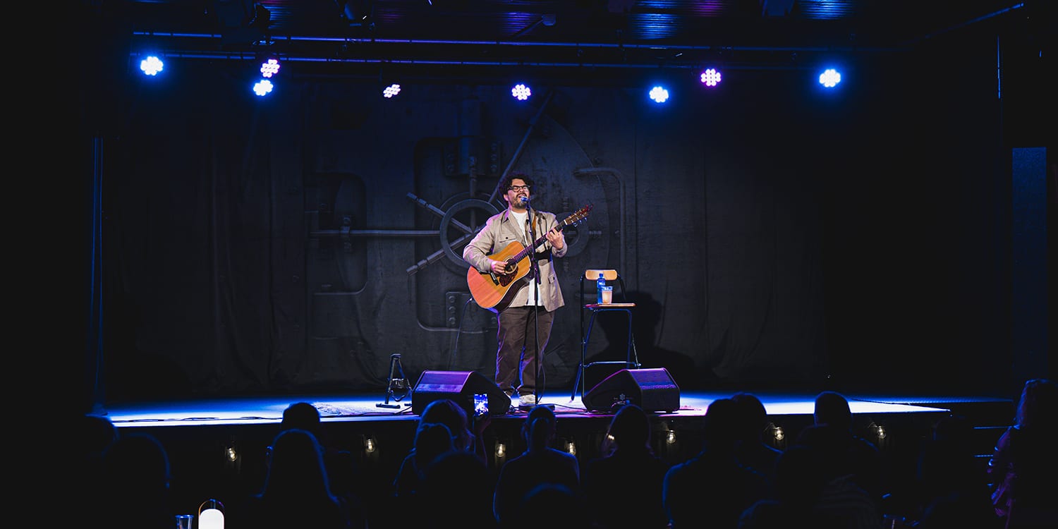 En musiker med briller og skjorte står på en scene og spiller akustisk gitar under blått scenelys. Bak ham er et bakteppe med et motiv av en stor bankhvelvdør. Publikum er synlige som mørke silhuetter i forgrunnen.