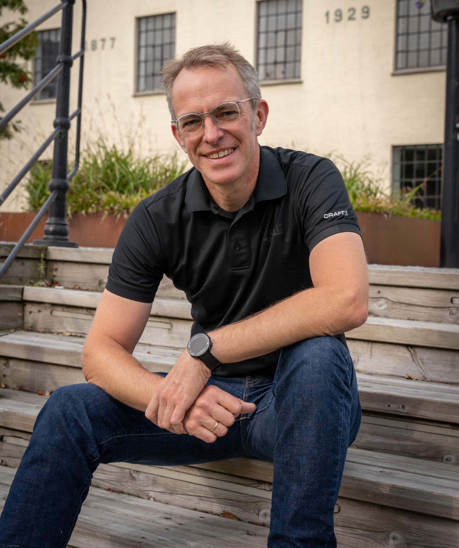 En mann med briller og kort hår sitter på en tretrapp foran en hvit murbygning merket "BRYNE MØLLE". Han smiler til kameraet, har armene hvilende på knærne og bærer en svart polo-skjorte og en smartklokke.