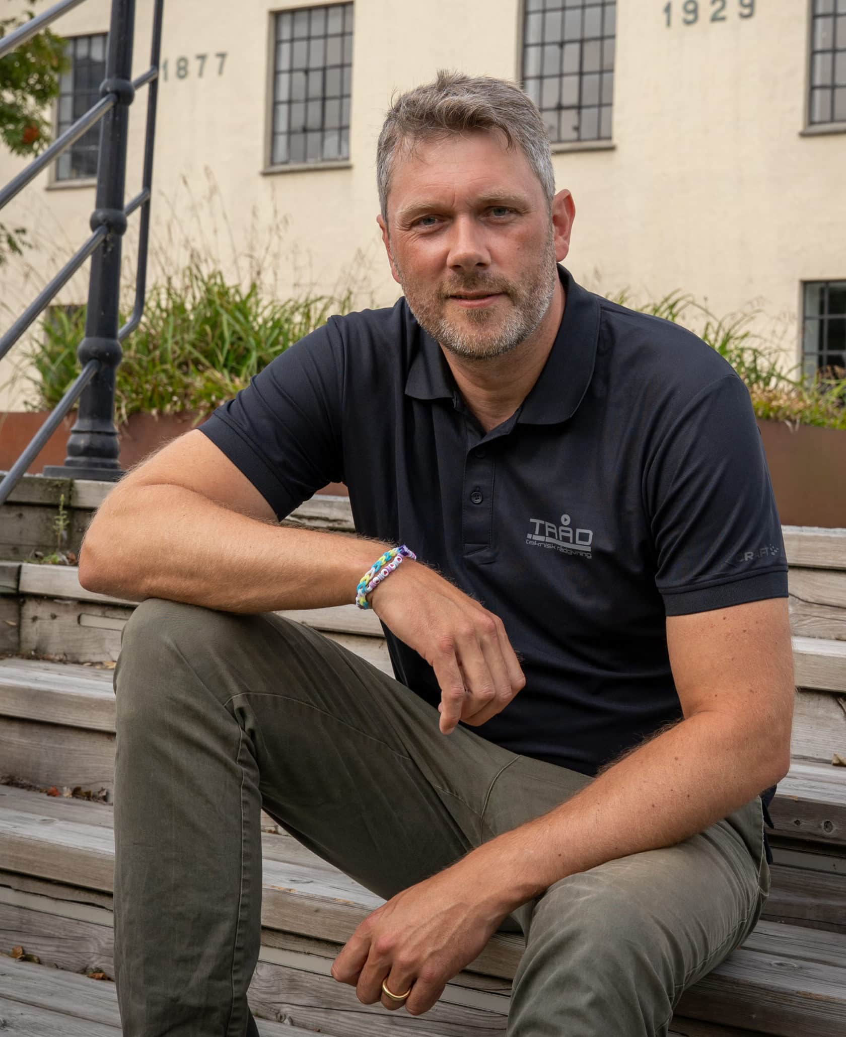 En mann med kort, gråsprengt hår og skjegg sitter på en tretrapp utendørs. Han er iført en svart polo-skjorte med "TRÅD"-logo og har et fargerikt perlearmbånd på håndleddet. En hvit murbygning med årstallet 1877 ses i bakgrunnen.