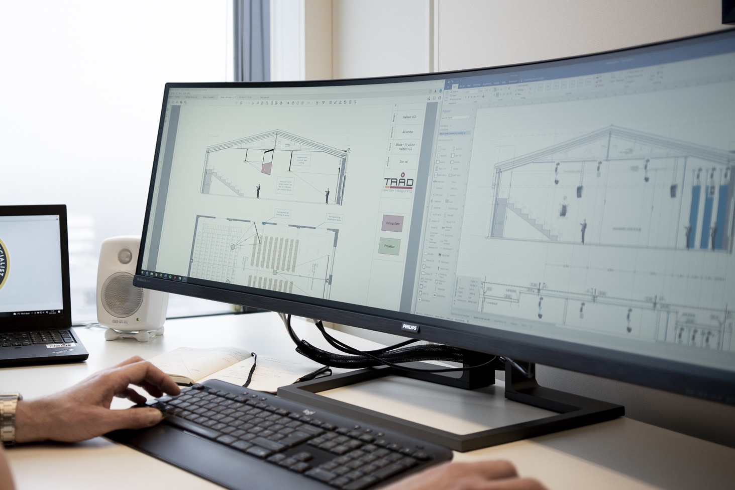 Nærbilde av en arbeidsstasjon med en ultrabred, buet dataskjerm som viser tekniske tegninger av et rom med AV-utstyr. En hånd hviler på et tastatur i forgrunnen, og en hvit Genelec-høyttaler står ved siden av skjermen.