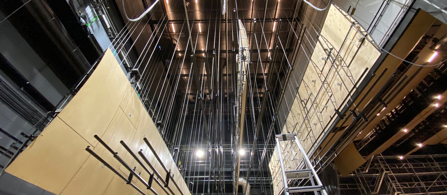 Et teknisk bilde tatt nedenfra og opp mot snoreloftet i et teater. En mengde vertikale wirer og trekkstenger henger fra taket. På sidene henger store, lyse kryssfinerplater (akustiske reflektorer) som er montert i riggsystemet.