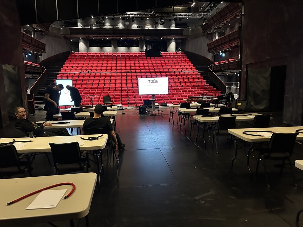Interiøret i en teatersal sett fra scenen. I forgrunnen står flere hvite skolepulter med svarte stoler, der det ligger papirer og kabler. En skjerm i midten viser logoen til "TRAD". Bak pultene stiger et stort amfi med røde seter oppover.