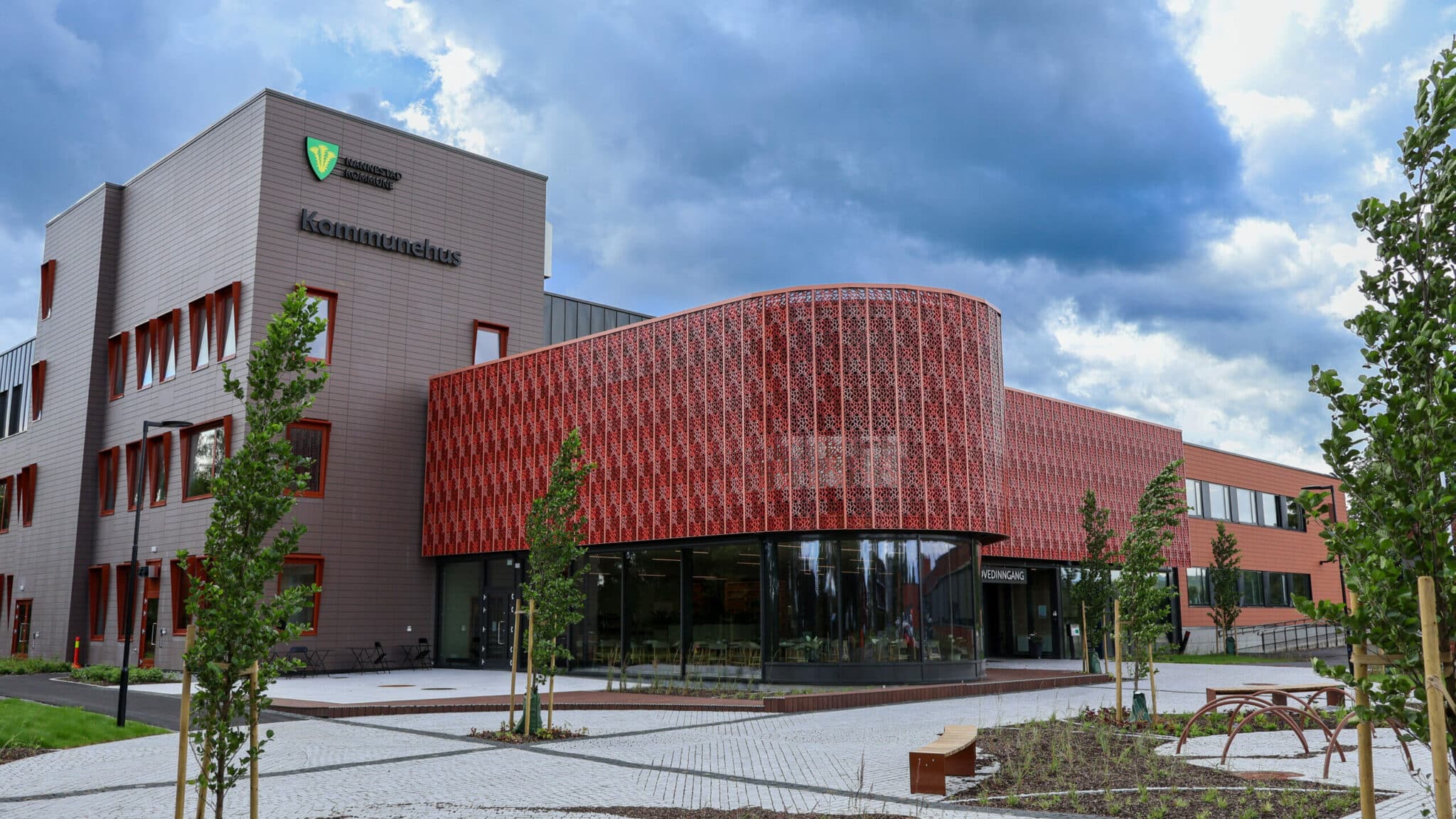 Eksteriøret til Nannestad Kommunehus. Bygget har en moderne kombinasjon av mørkebrun kledning og en iøynefallende buet fasade dekket av et rødbrunt, perforert metallmønster. Foran inngangen er det en steinlagt plass med unge trær og benker.
