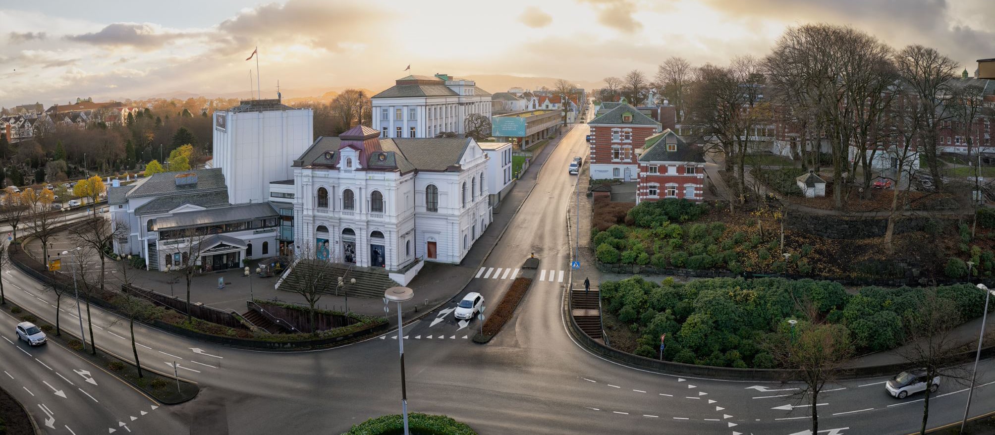 Et oversiktsbilde i vidvinkel av Rogaland Teater og de omkringliggende gatene i Stavanger under en lav ettermiddagssol. Den historiske teaterbygningen i hvit mur dominerer krysset, omgitt av grøntarealer, nakne trær og moderne infrastruktur.