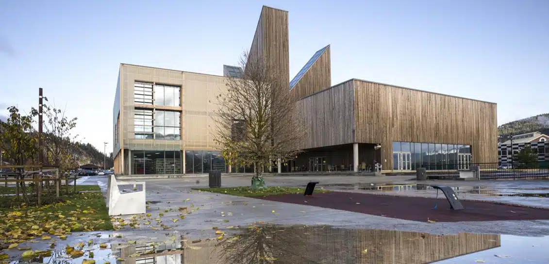 Eksteriøret til et moderne kulturbygg med vertikal trekledning. Bygget har særpreget arkitektur med høye, spisse takformer som minner om katedralspir. En åpen plass med vanndammer og høstløv ligger i forgrunnen.