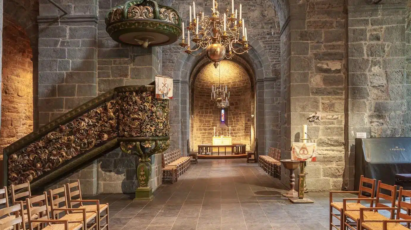 Interiøret i en historisk steinkirke (Stavanger domkirke). En rikt utskåret og fargerik prekestol i barokkstil dominerer venstre side. En stor messingkrone henger fra taket i midtgangen som fører mot alteret i bakgrunnen.