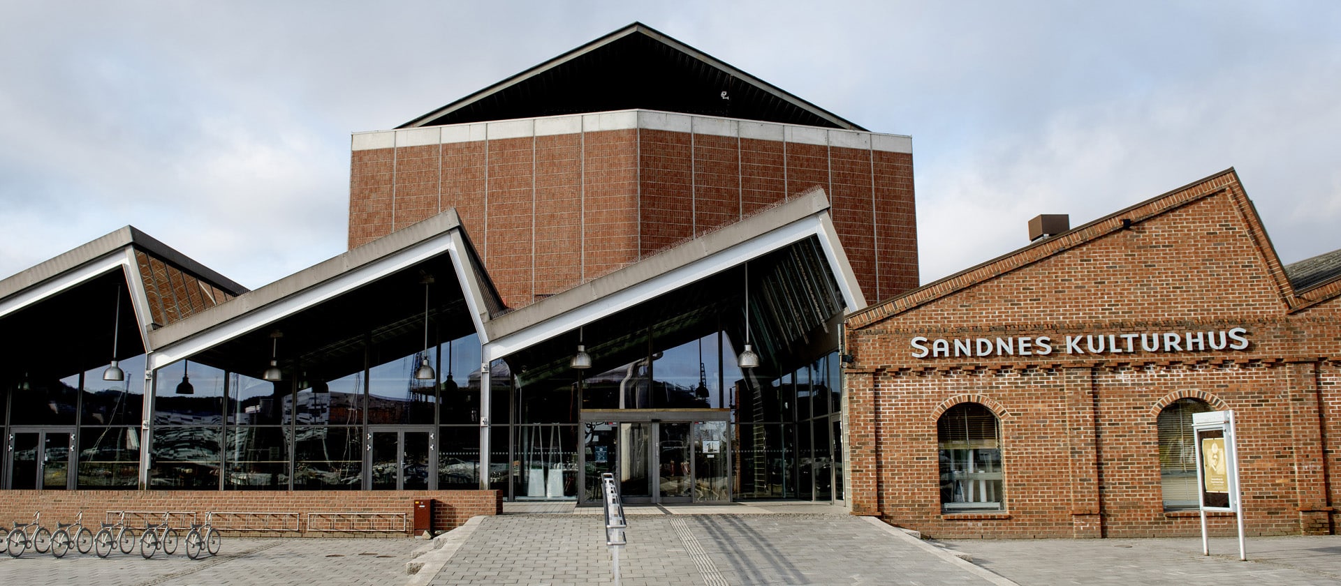 Eksteriøret til Sandnes Kulturhus på en dag med lett skydekke. Bygget er i rød murstein med store glassflater ved inngangspartiet og et karakteristisk sagtann-formet tak over den moderne delen.