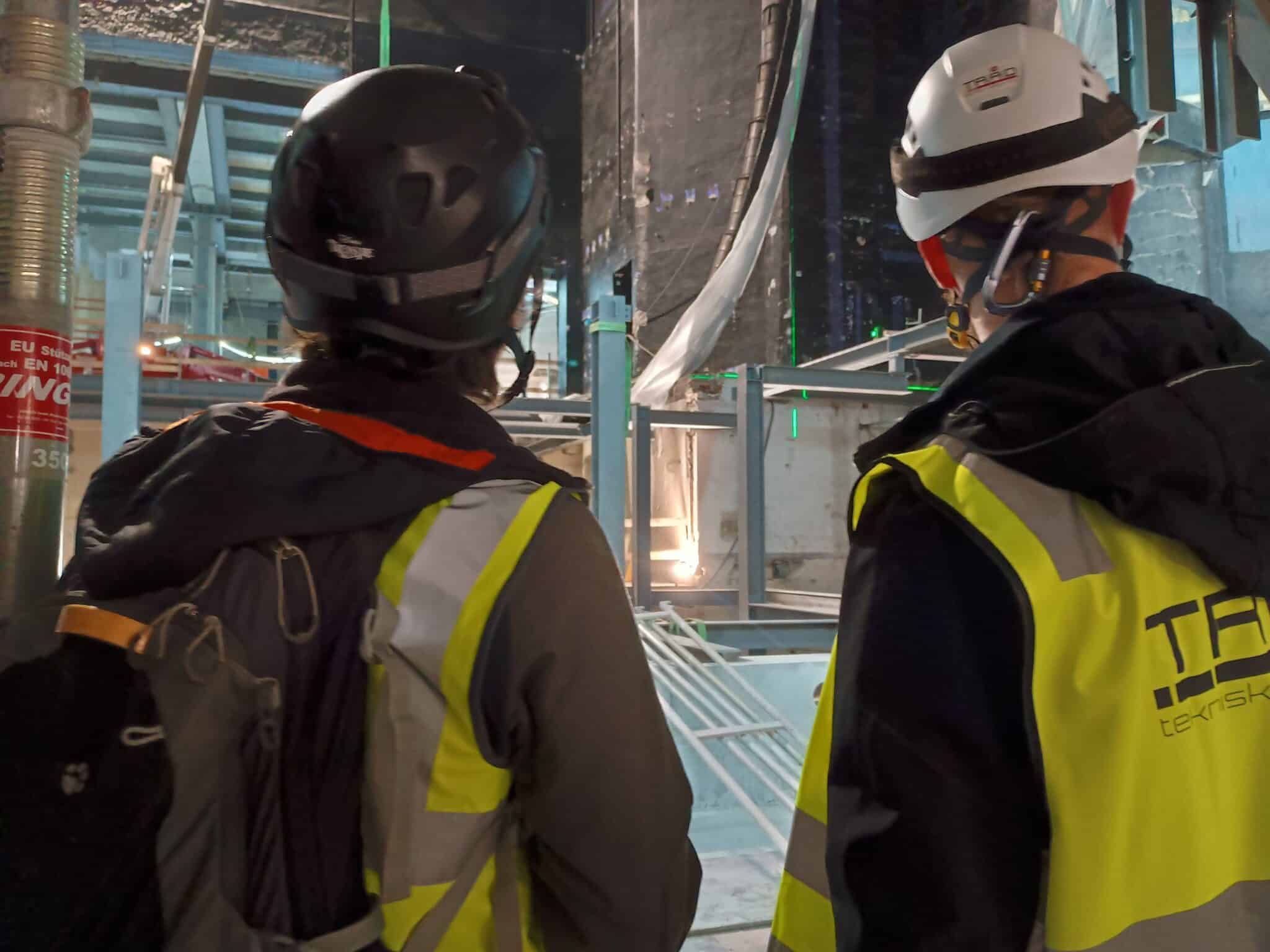 To personer i gule refleksvester og vernehjelmer sett bakfra på en byggeplass. De observerer tekniske installasjoner i et stort, åpent rom med betongsøyler og stålbjelker.