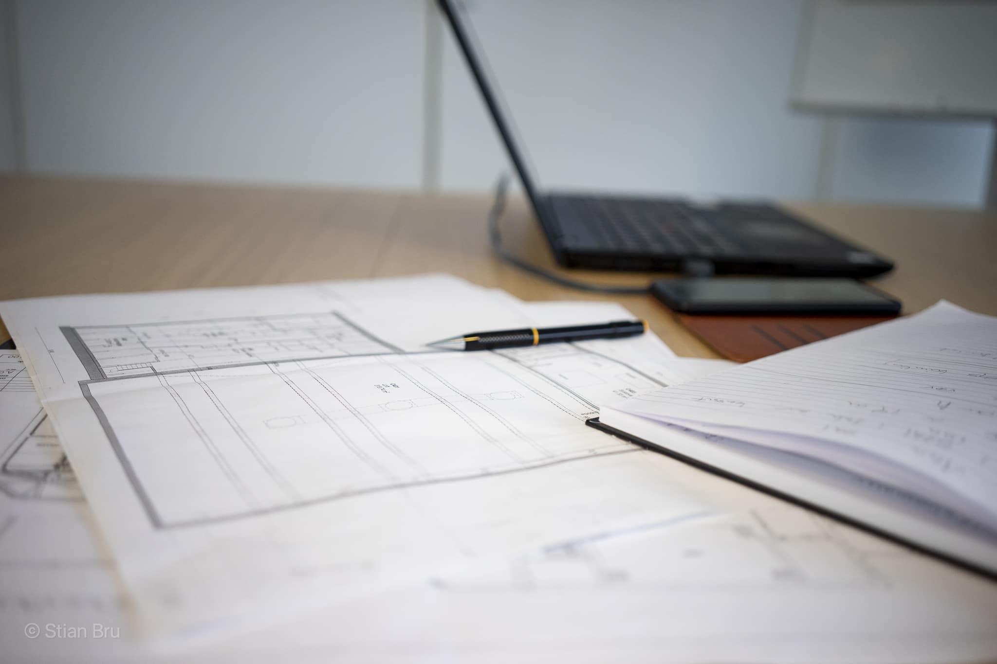 Tekniske tegninger og plantegninger ligger spredt utover et lyst trebord. En svart penn ligger oppå papirene, og en bærbar datamaskin er delvis synlig i bakgrunnen.