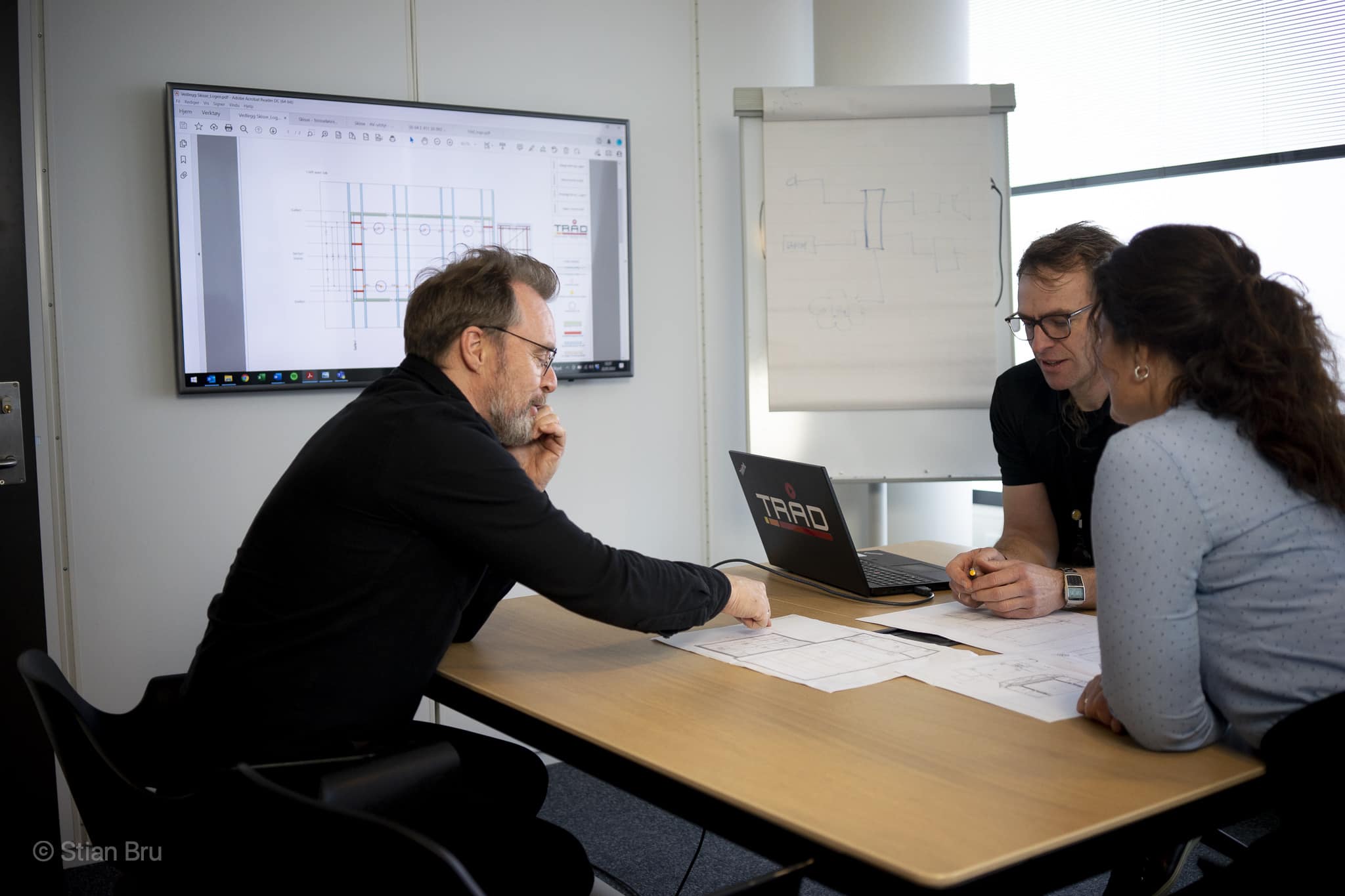 Tre personer sitter rundt et møtebord og diskuterer tekniske tegninger. En stor skjerm på veggen viser en detaljert plantegning av et rom med tekniske spesifikasjoner.