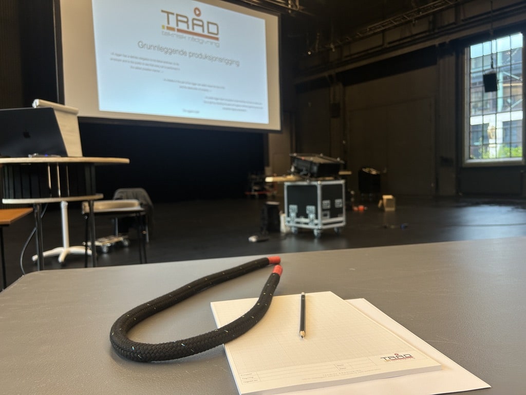 Et nærbilde av en hvit blokk med en blyant og et svart tau på et bord i et mørkt lokale. I bakgrunnen ses et lerret som viser en presentasjon fra "TRÅD teknisk rådgivning" om grunnleggende produksjonsrigging.